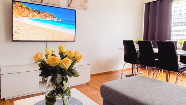 A living room with a flat-screen TV, a coffee table with a vase of yellow roses, grey sofas, and a dining area with black chairs.