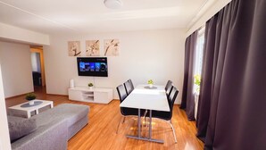 A modern living room with a white sofa, a dining table with chairs, a flat-screen TV mounted on the wall, and a window with dark curtains.