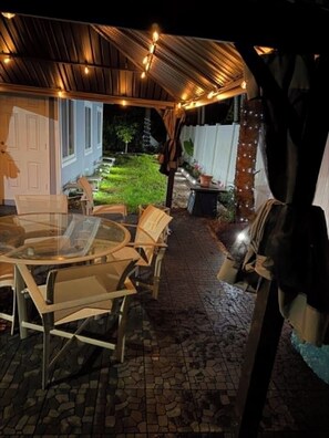 Un'area patio esterna con tavolo e sedie in vetro, luci decorative a catena e pavimento in mattoni.