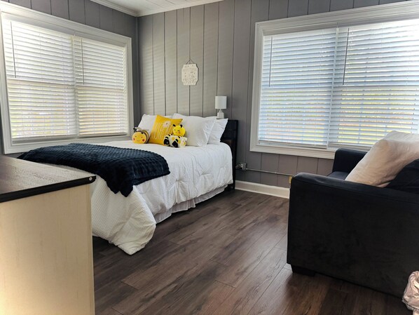 A bedroom with a bed, a bedside table, a chair, and a window with blinds. - Cottage home near downtown Douglasville’s Greystone Amphitheater. (Douglasville)