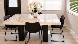 Un comedor con una mesa de mármol, sillas negras, un florero con flores blancas, un reloj de pared y una ventana con persianas.