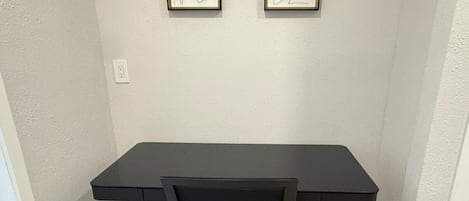 Un bureau noir avec une chaise en tissu grillagé, dans une pièce aux murs de couleur claire et au plancher en bois. Deux œuvres d’art encadrées sont accrochées au mur au-dessus du bureau.