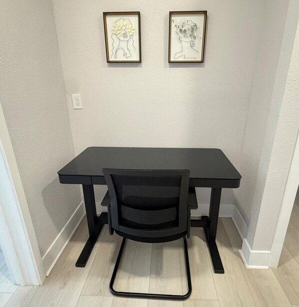 Un bureau noir avec une chaise en tissu grillagé, dans une pièce aux murs de couleur claire et au plancher en bois. Deux œuvres d’art encadrées sont accrochées au mur au-dessus du bureau.
