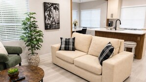 Una sala de estar moderna con un sofá color beige, sillones verdes, una mesa de centro, un ventilador de techo y una cocina al fondo.