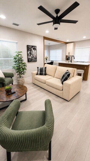 Un soggiorno moderno con un divano color beige, poltrone verdi, un tavolino da caffè, una ventola a soffitto e una cucina sullo sfondo.