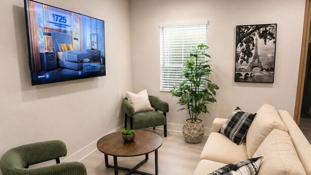 Una sala de estar con un sofá beige, dos sillones, una mesa de centro, un televisor montado en la pared, una ventana con persianas, una planta en maceta y un cuadro enmarcado.