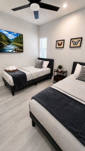 Une chambre avec deux lits, un ventilateur de plafond, un tableau représentant une rivière et deux papillons encadrés.