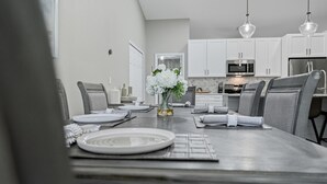 A modern dining area with a long table set for four, surrounded by grey upholstered chairs. A vase with white flowers is placed in the center. - Newly Remodeled, Spacious 4-Bedroom Getaway (Palm Coast)