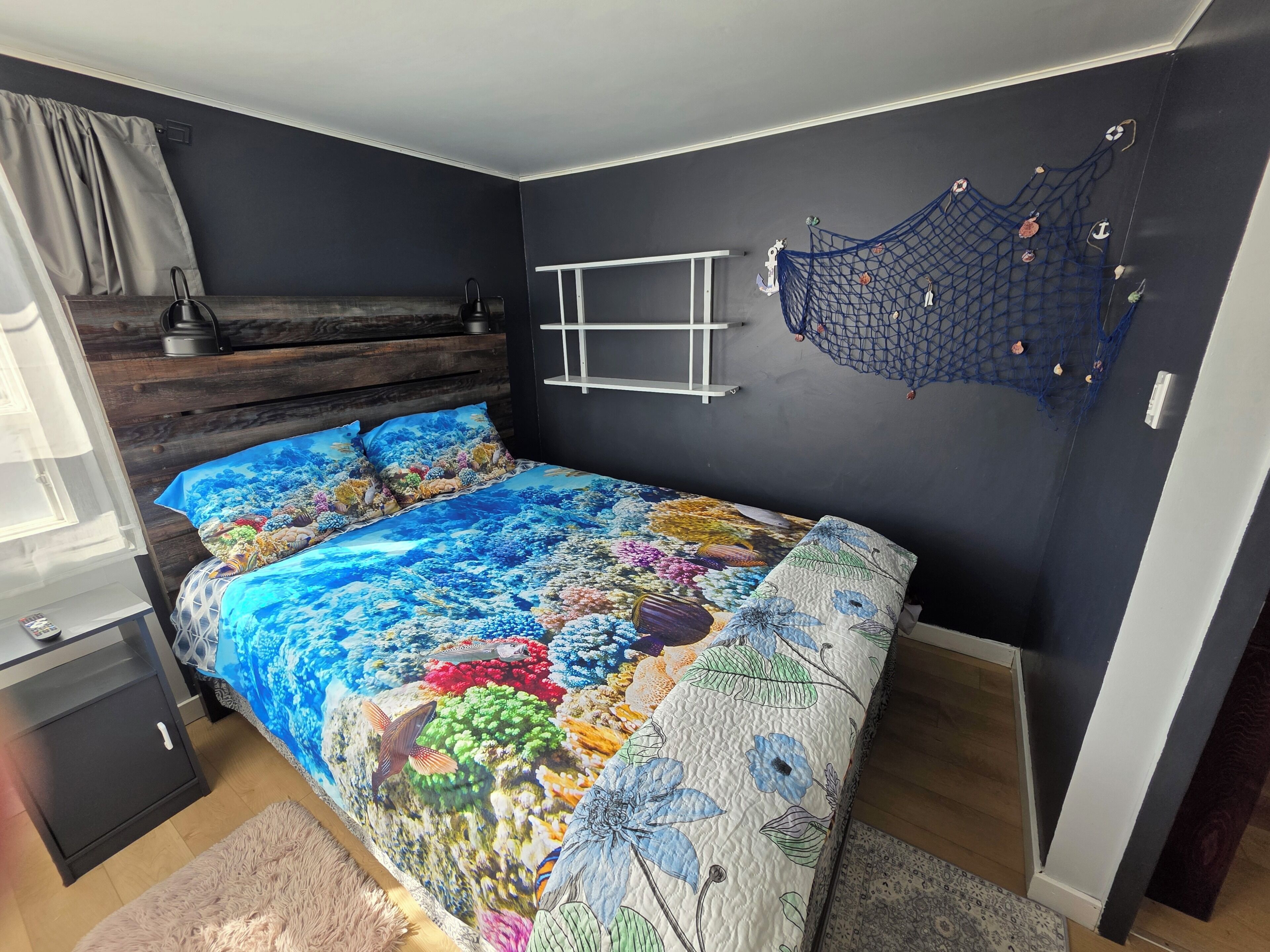 Un dormitorio con una cama, cabecera de madera, estante en la pared, una hamaca y una ventana con cortinas.