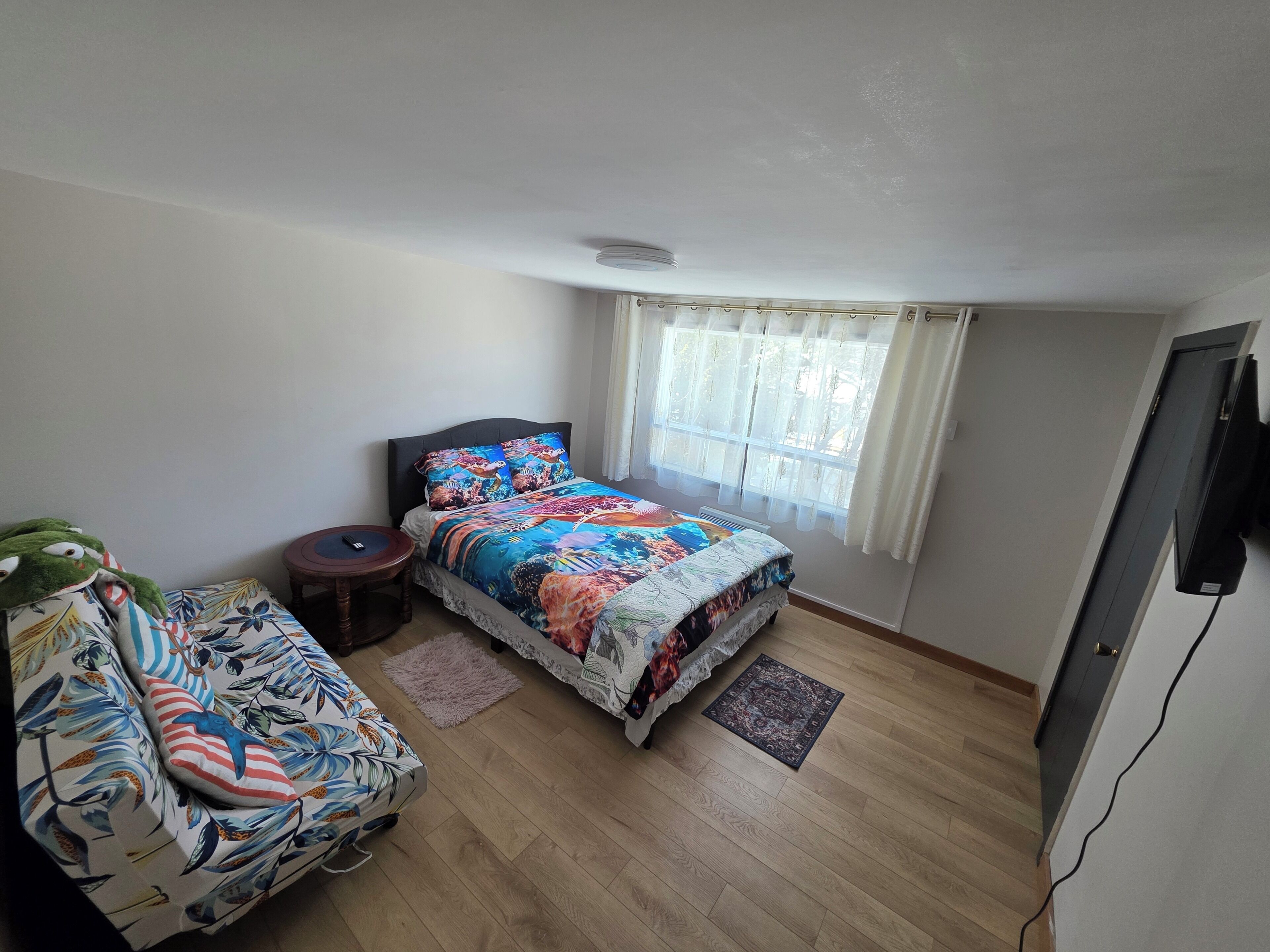 A bedroom with a bed, a small table, a sofa, a window with curtains, and a flat-screen TV.