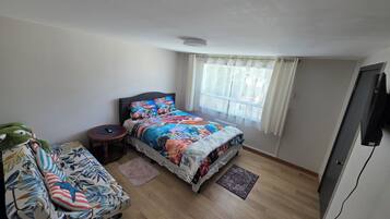 A bedroom with a bed, a small table, a sofa, a window with curtains, and a flat-screen TV.