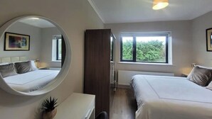 Une chambre avec deux lits, un miroir rond, un bureau et une fenêtre donnant sur un paysage verdoyant.