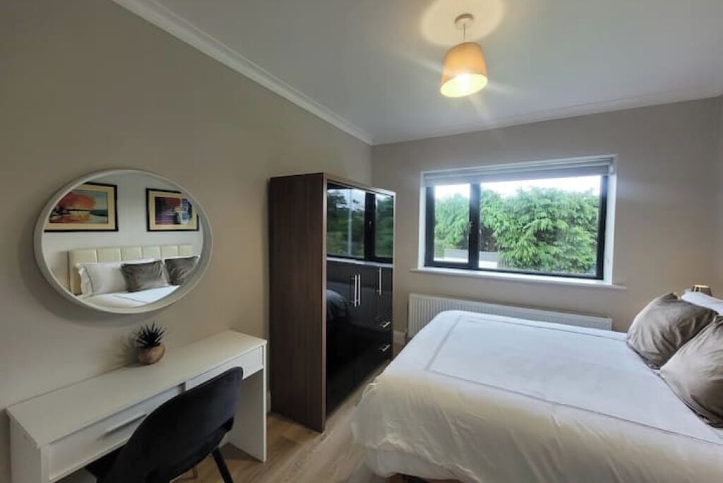 A bedroom with a bed, a desk, a chair, a wardrobe, a round mirror, and a window with a view of greenery.
