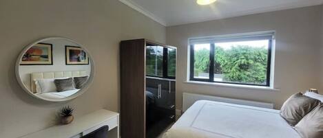 A bedroom with a bed, a desk, a chair, a wardrobe, a round mirror, and a window with a view of greenery.
