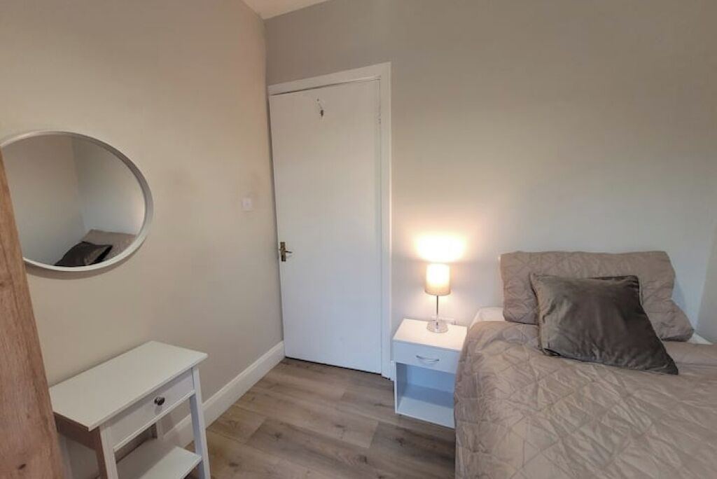 Una camera da letto con un letto, un comodino con una lampada, un piccolo mobiletto bianco, uno specchio rotondo e una porta.