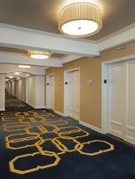 Un pasillo de hotel con alfombra estampada, varias puertas y una lámpara decorativa en el techo.