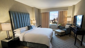 A hotel room with a large bed, a desk with a lamp, a television, and a view of a cityscape through the window. - The Marc Hotel (Milwaukee)