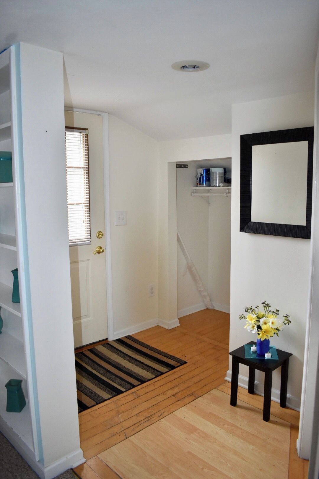 Un couloir avec un plancher en bois, une porte donnant sur une pièce, une petite table sur laquelle est posé un vase de fleurs et un miroir fixé au mur.