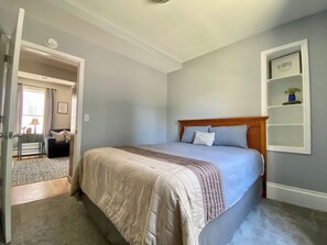 A bedroom with a bed, a headboard, a bedside table, and a wall-mounted shelf with a framed item. - Cozy Main Street Flat (Adrian)