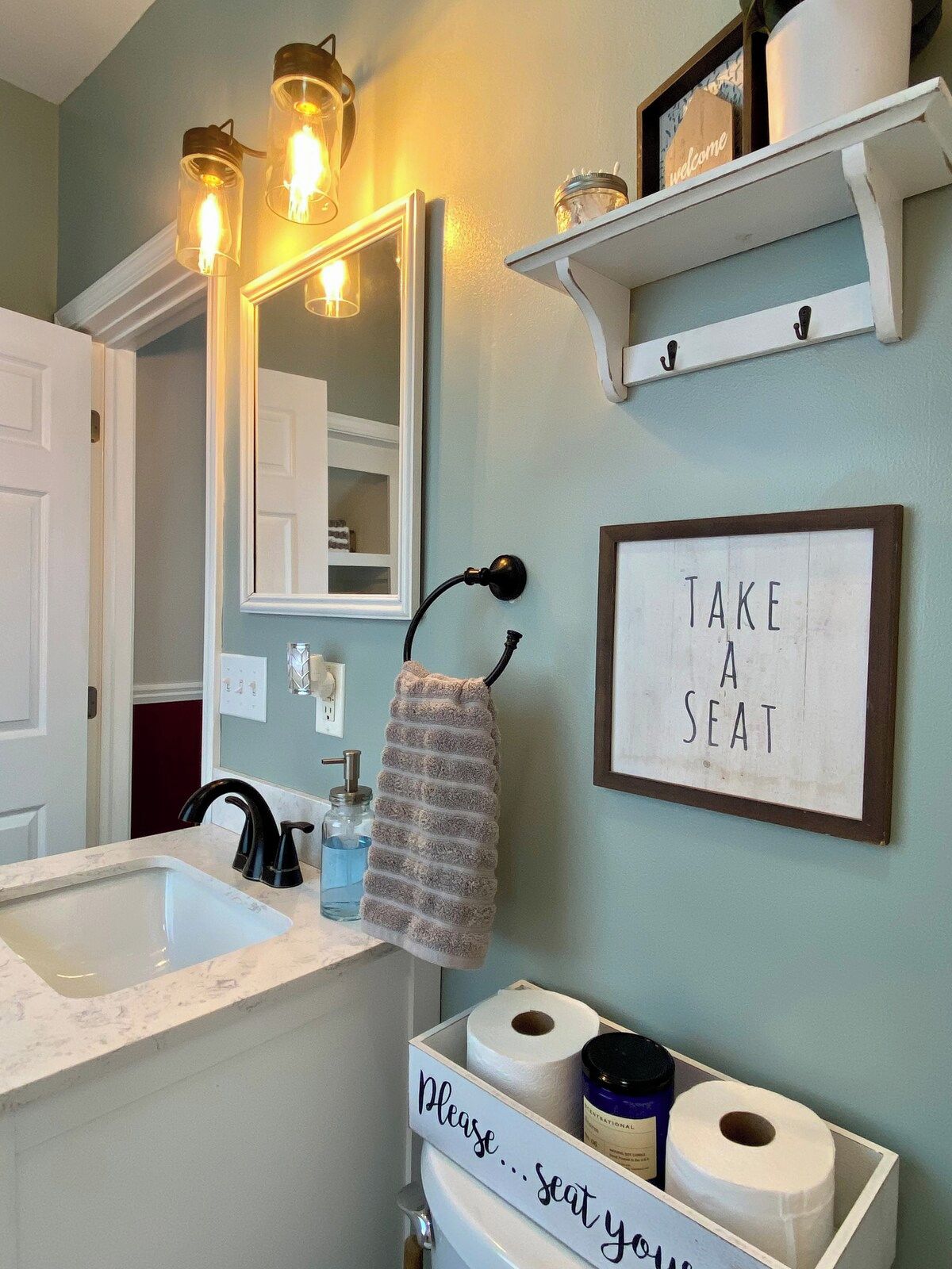 A bathroom with a sink, mirror, towel rack, and a sign that says 'TAKE A SEAT'.