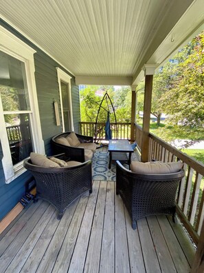 A covered porch with wicker furniture, a swing, and a view of a green landscape. - Cozy Main Street Flat (Adrian)