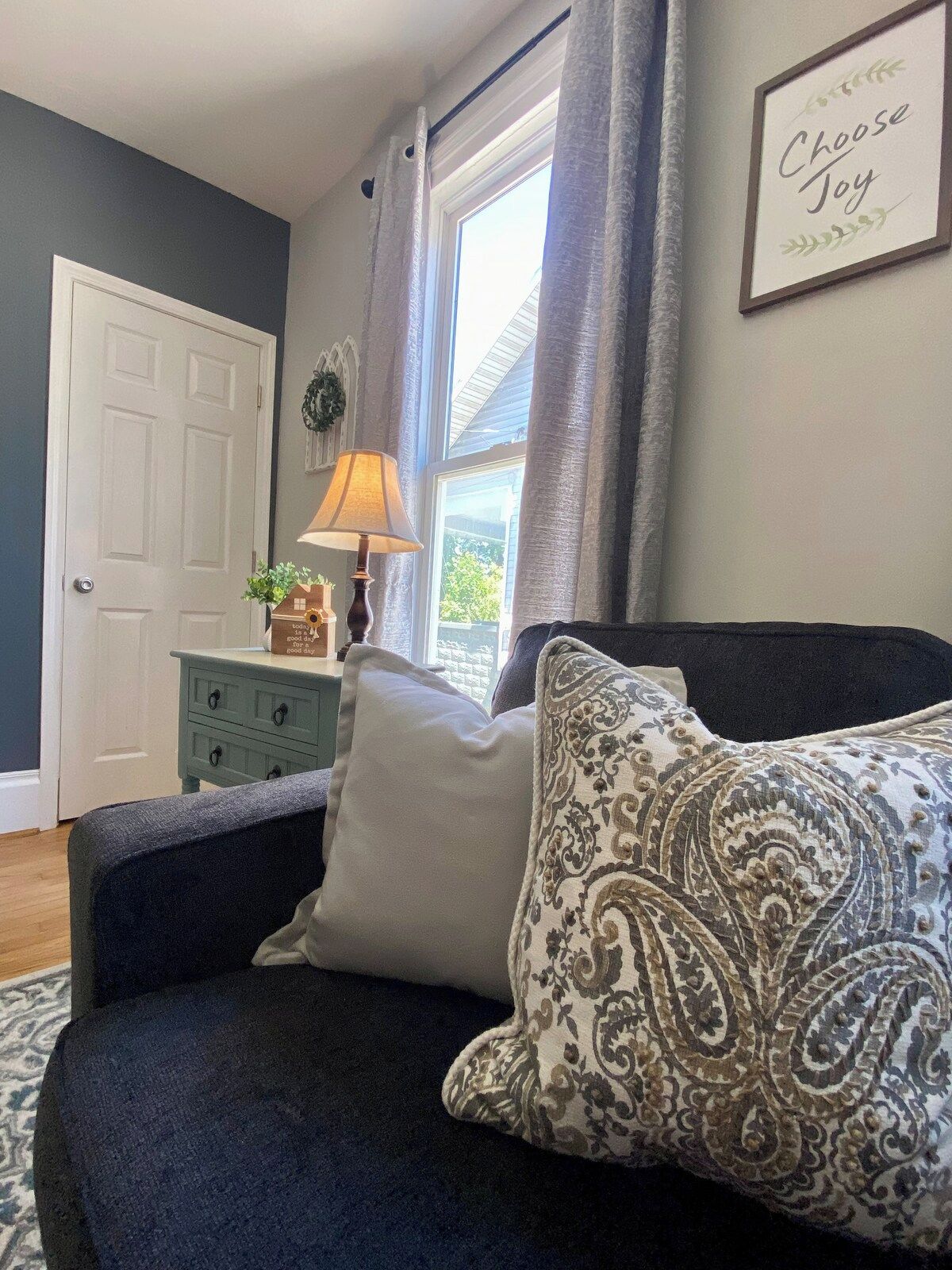 Une chambre avec un canapé bleu foncé, un coussin décoratif à motifs, un coussin blanc, une petite table d’appoint en bois avec une lampe, et un panneau mural sur lequel on peut lire : « Choisissez la joie ».