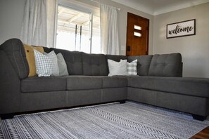 A grey sectional sofa with multiple pillows, a patterned rug, and a 'welcome' sign on the wall. - Cozy Garden House (Adrian)