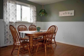 A dining room with a wooden table and chairs, a window with patterned curtains, and a hanging plant. - Cozy Garden House (Adrian)