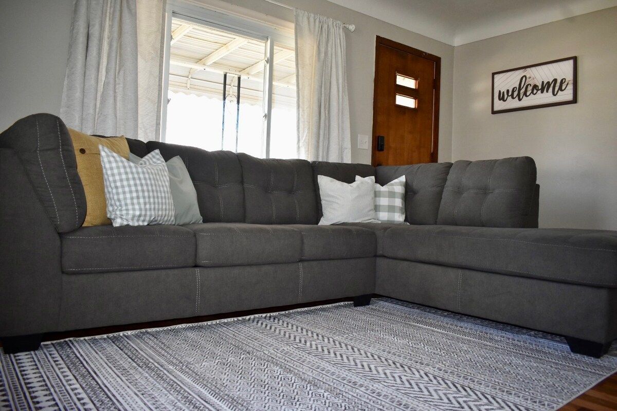 A grey sectional sofa with multiple pillows, a patterned rug, and a 'welcome' sign on the wall.