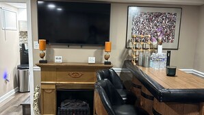 A modern living room with a fireplace, a large flat-screen TV, a bar area with stools, and a framed photo on the wall. - Cozy private one bedroom basement apartment with private parking and entrance. (Landover)