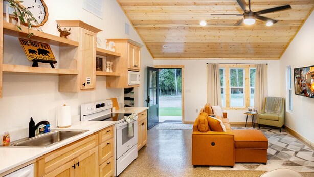 A modern kitchen with wooden cabinets, a microwave, and a sink. There is a dining area with a table and chairs, and a sofa. The room has a wooden ceiling and a large window. - Private Cabin Villege with Hot Tubs & Firepit for 16 people (Cherokee County)