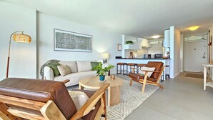 A modern living room with a sofa, coffee table, and chairs, a kitchen in the background, and a dining area with stools.
