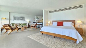 A modern hotel room with a large bed, a sofa, a dining area, and a kitchen in the background.