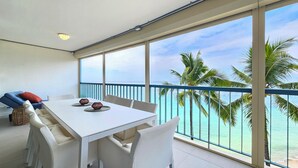 A balcony with a white table and chairs, overlooking a turquoise sea and palm trees.