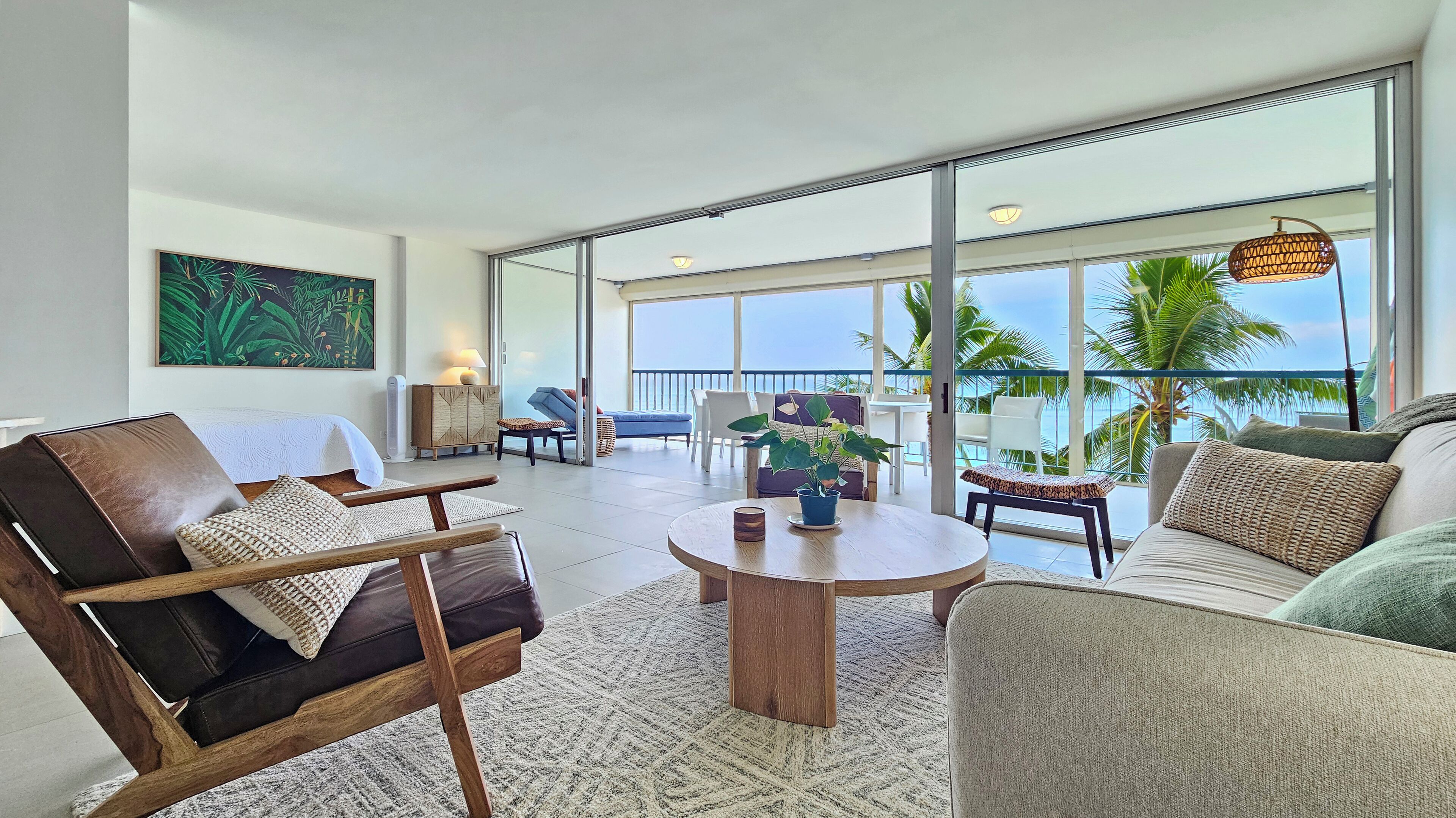 A spacious living room with a large window overlooking a balcony with a view of palm trees and the ocean. There are several seating areas, including a sofa, armchairs, and a coffee table.
