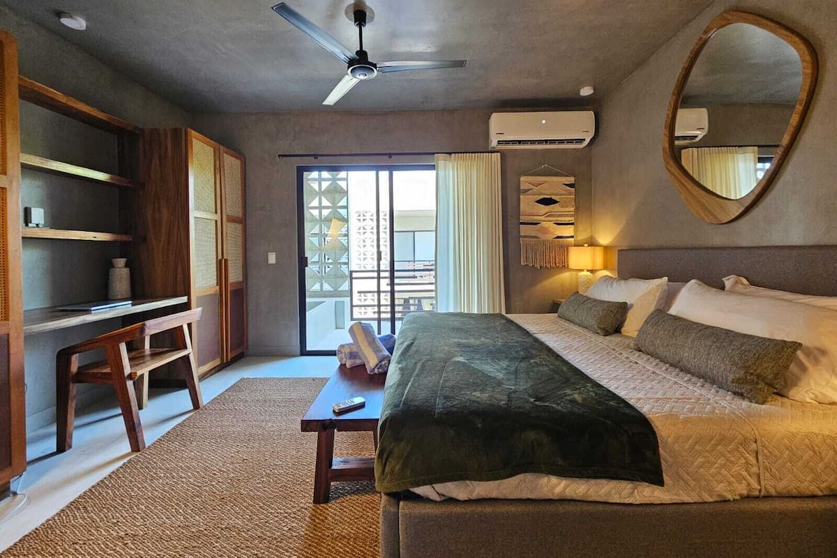 A hotel room with a bed, a desk, a chair, a ceiling fan, an air conditioning unit, a mirror, and a view through a sliding door.