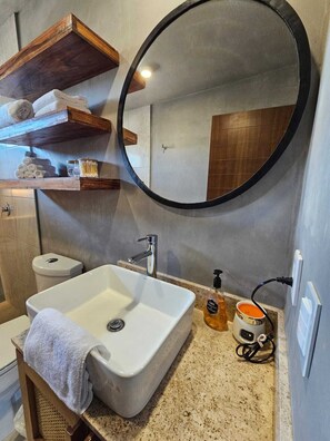 Ein Badezimmer mit Waschbecken in Weiß, einem runden Spiegel, Holzregalen mit Handtüchern, einer Toilette und einer Kaffeemaschine.