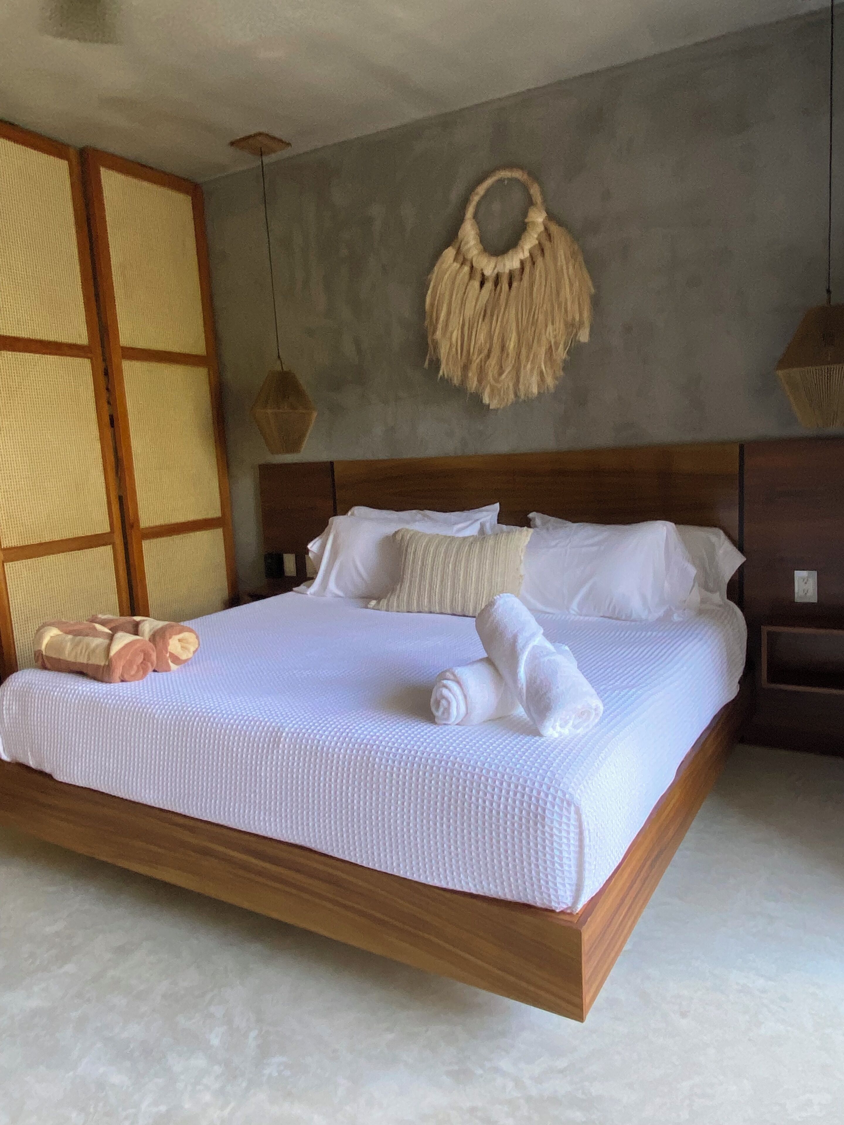 Ein Bett mit weißer, gemusterter Bettdecke, einem Holz-Kopfschild und einem gewebten Wandbehang aus Makramee.