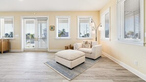 A spacious living room with a beige sofa, ottoman, and a floor lamp. There are large windows with blinds, a door leading outside, and a small side table with a lamp.