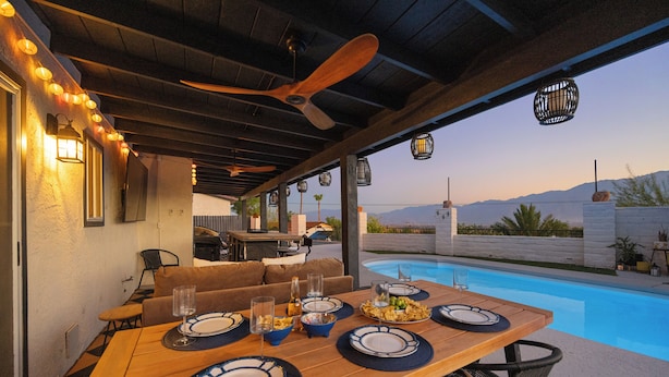 An outdoor dining area with a pool, wooden table, chairs, and a ceiling fan.