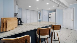 A kitchen with a granite countertop, wooden chairs, a coffee maker, and a refrigerator. - 🐚Seashell Oasis B-1 - Beautiful Condo steps to the Beach! (Redington Shores)