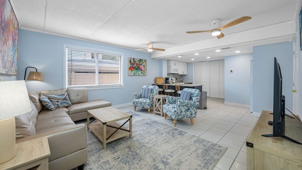 A spacious living area with a sofa, coffee table, and patterned chairs. A television is on a wooden stand, and a ceiling fan is visible. - 🐚Seashell Oasis B-1 - Beautiful Condo steps to the Beach! (Redington Shores)