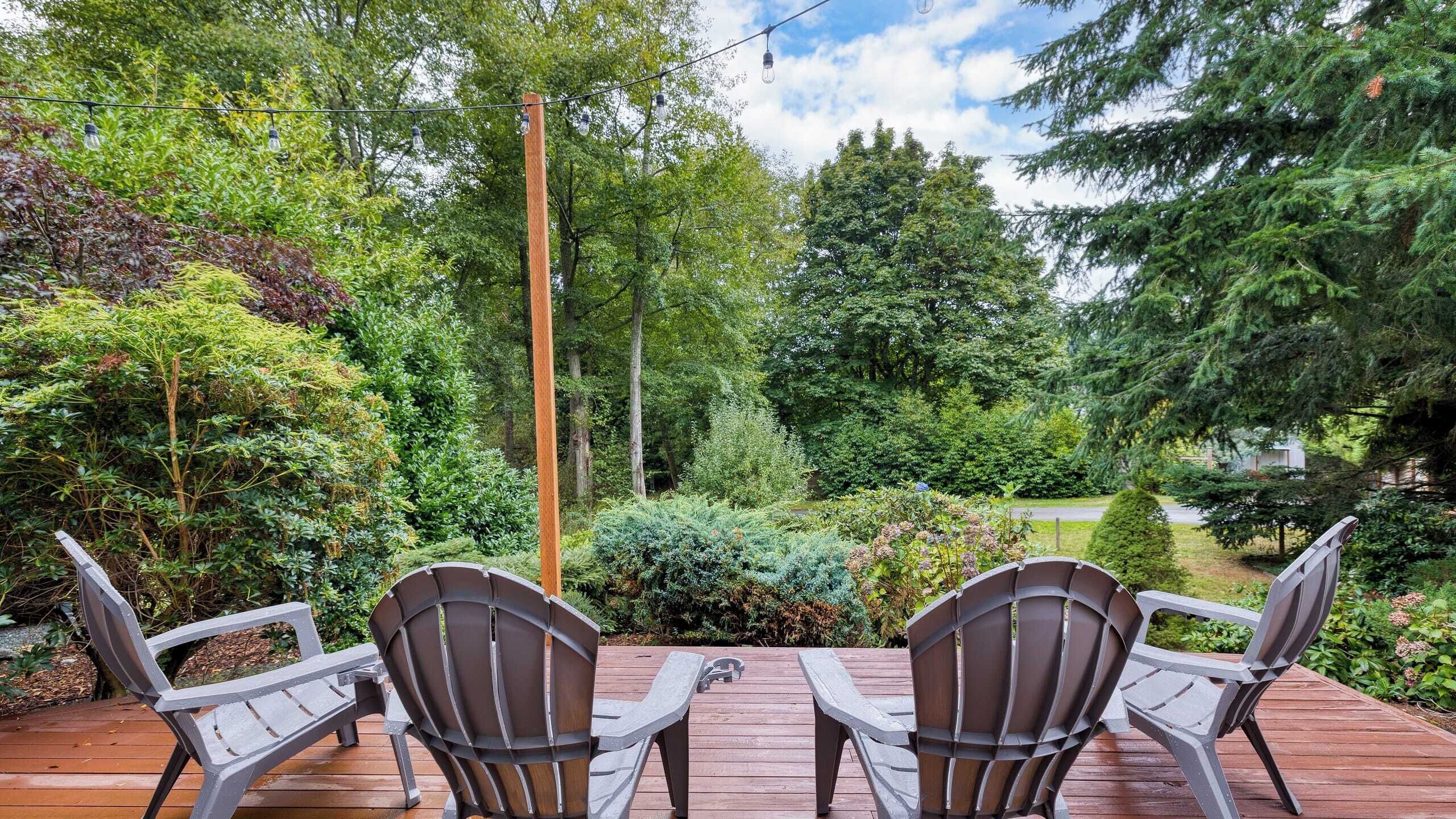 Una terraza de madera con cuatro sillas Adirondack mirando un jardín exuberante con diversas plantas y árboles.
