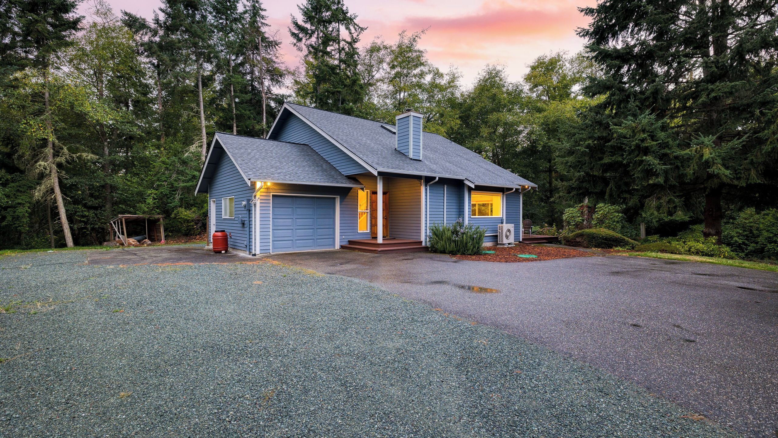 A two-story house with a garage, surrounded by a gravel driveway and a forested area.