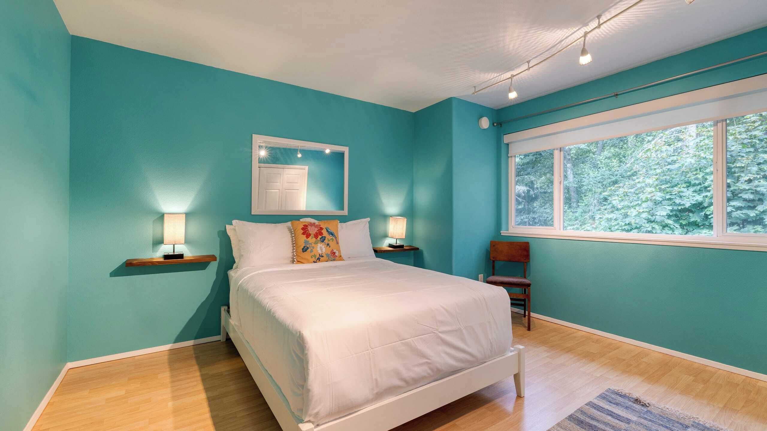 A bedroom with teal walls, a white bed, two bedside tables with lamps, a window with a view of greenery, and a wooden floor.