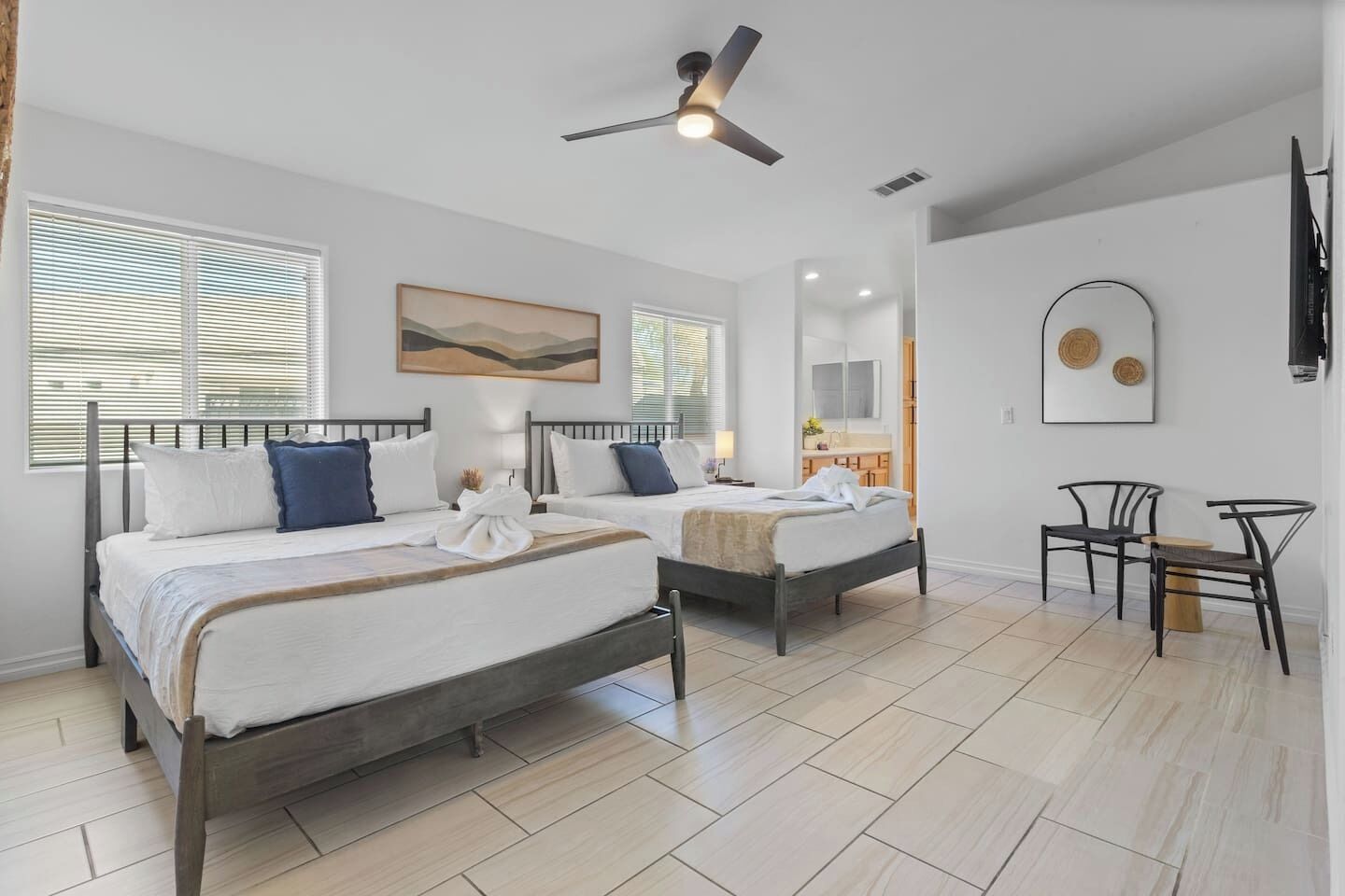A hotel room with two beds, a ceiling fan, a bathroom with a mirror, and a small seating area.