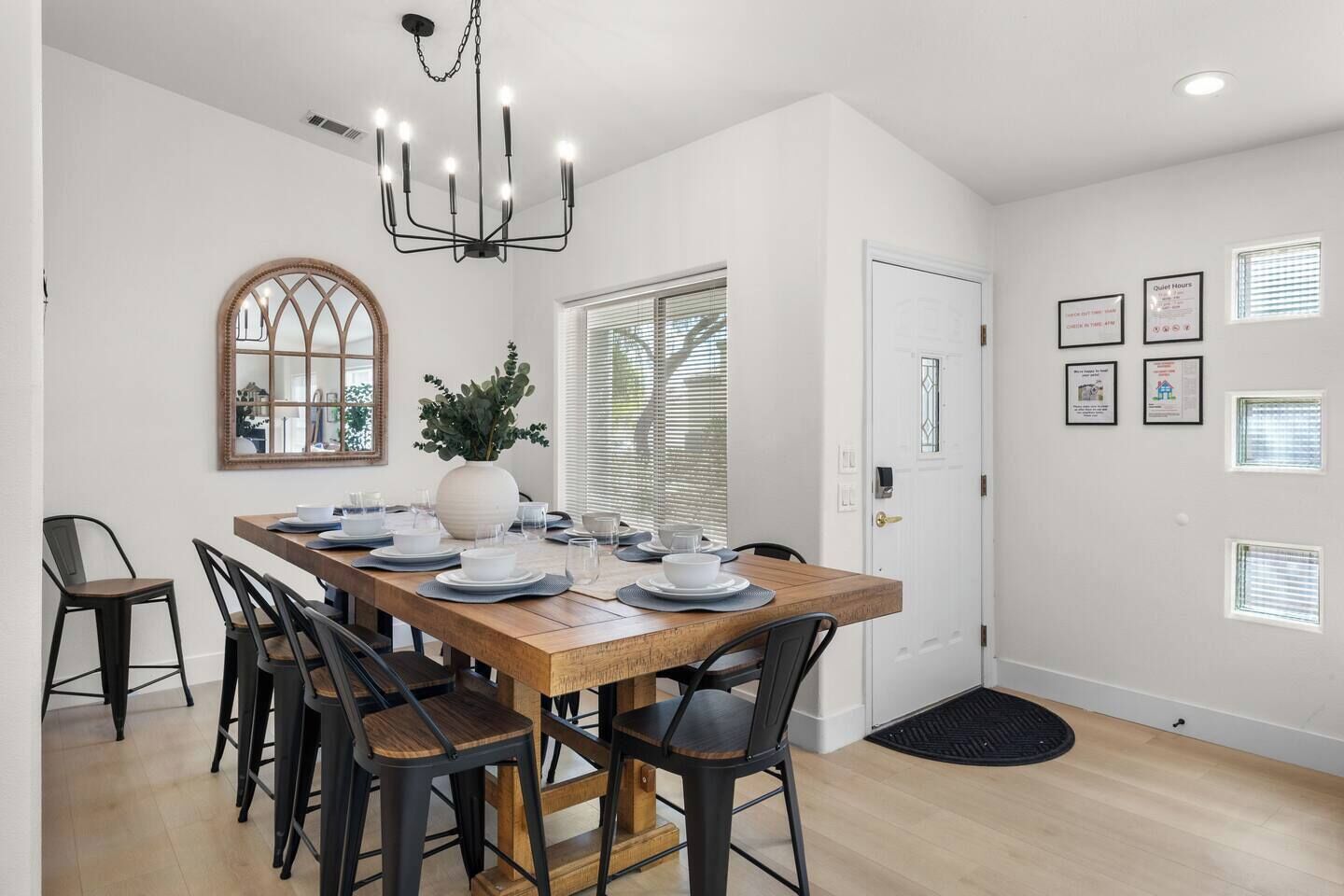 A dining area with a wooden table set for six, black chairs, a chandelier, a large mirror, and a potted plant.