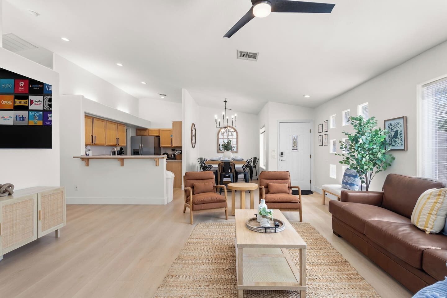A modern living room with a brown leather sofa, wooden coffee table, and two armchairs. A kitchen with wooden cabinets and a dining area with a chandelier are visible in the background.