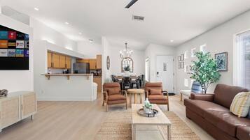 A modern living room with a brown leather sofa, wooden coffee table, and two armchairs. A kitchen with wooden cabinets and a dining area with a chandelier are visible in the background.