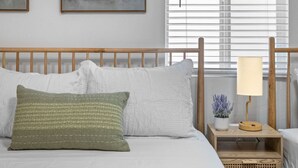 Una cama bien tendida con almohadas blancas y verdes, un cabecero de madera y una mesita de noche con una lámpara y una planta pequeña.